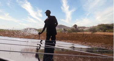 A person works on an irrigation system in a field. There are solar panels in the foreground and hills, bushes, and trees in the background.