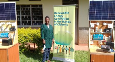 On the grass in front of a building, Christine Alobo stands next to a roll-up banner that reads "Renewable Energy Generations in the Hands of Citizens". She is wearing a green lab coat and smiling. At her side are instruments and photovoltaic kits.