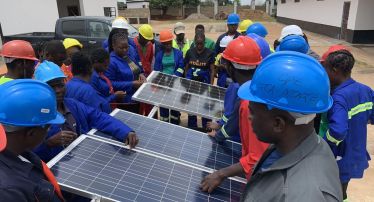 A group of young adults stands around some photovoltaic panels. They are wearing overalls of different colours. Some of them are also wearing helmets.