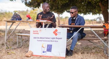 Two people are sitting behind tables with a tree in the background. A sign in front of them shows the logos of SunnyMoney and SolarAid, a lamp and a photovoltaic panel. It reads: Ask us about solar light repair. Trained technicians work here.