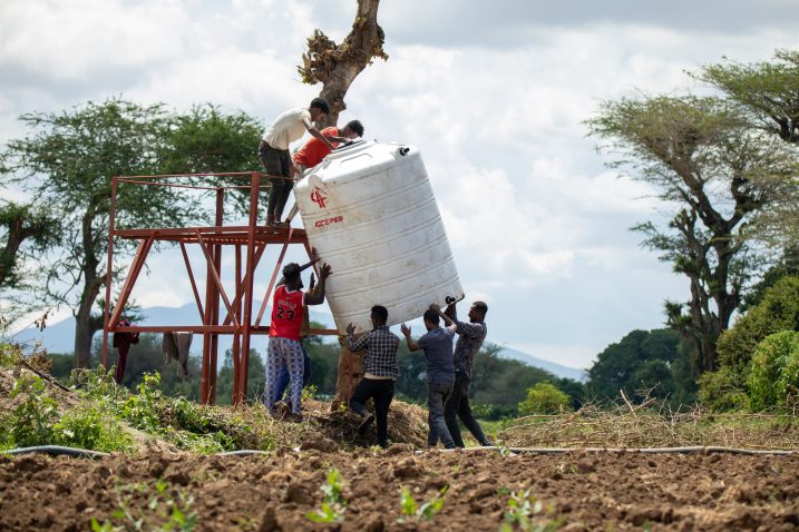 Six people lift a cistern onto a metal stand in a field, with trees and hedges in the background.