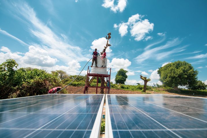 Solar panels in the foreground, two people stand on a metal frame with a cistern on top. Blue sky with white clouds, a field, and some trees in the background.