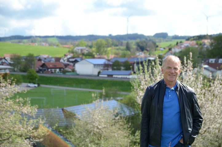 Günter Mögele, Deputy Mayor of Wildpoldsried, Bavaria, in front of some flowering hedges. In the background, a village with solar panels on many roofs and windmills on a hill behind the houses.