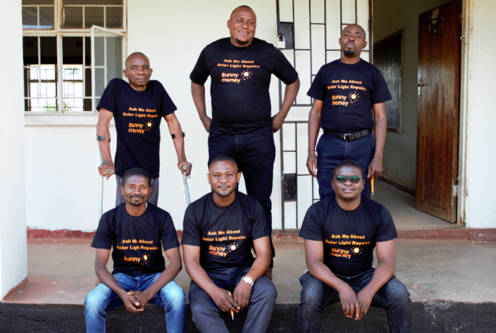 Six people stand on the veranda of a building. They are wearing T-shirts with the SunnyMoney logo and the words "Ask me about solar light repairs".