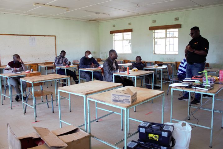 Six people are sitting at desks in a classroom. One person is standing and talking. There are tools and electrical equipment on the desks.