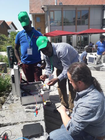 Outside a building, three people work with a photovoltaic kit in a suitcase and a meter.