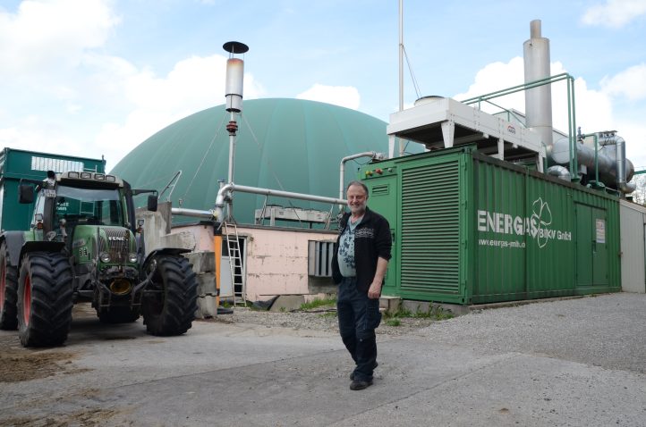 A person stands in front of a biogas plant. A tractor with a trailer is parked next to the plant.