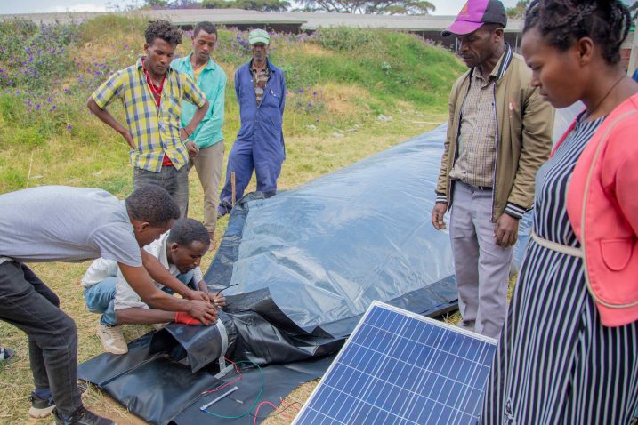 Seven people stand on a field, two of them are working on a construction with plastic foils and a solar panel. There are houses in the background.