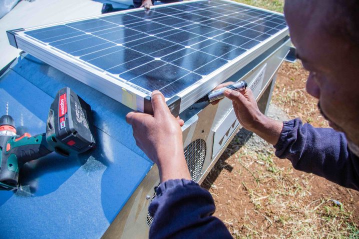 A person works with a tool on a solar dryer. A cordless screwdriver lies next to the solar panel.