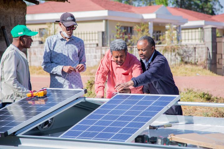 Four people are examining a solar dryer. There are houses in the background.