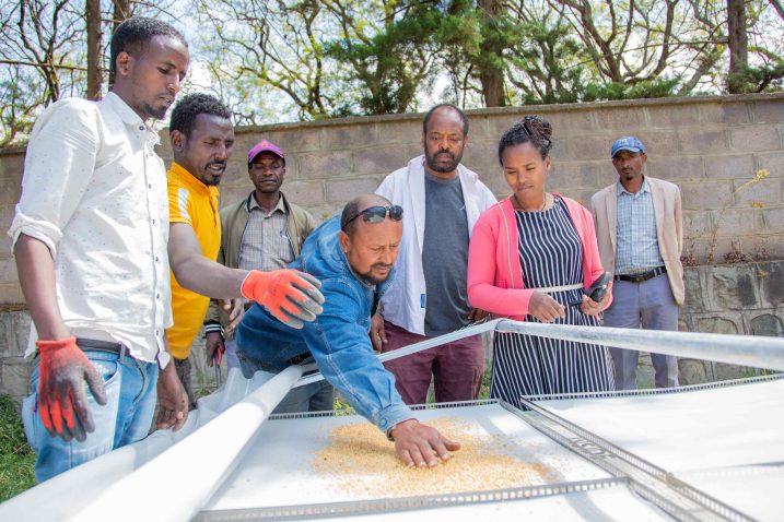 Seven people stand around a solar dryer. One person is placing seeds in the dryer.